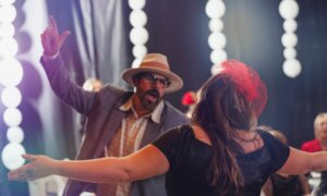 Man wearing a derby hat, suit, and glasses dancing with a woman wearing a black dress and red pill-box hat.