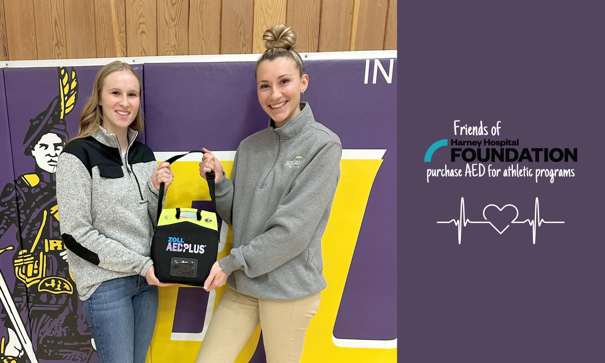 Tall, blonde teenage girl and tall, blonde young woman pose with an AED machine in the Burns High School gym.